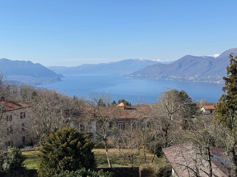 The image shows a scenic view of a lake surrounded by mountains. In the foreground, there are several buildings with red tile roofs, and the landscape is dotted with trees. The lake appears to be calm and serene, reflecting the surrounding mountains and s