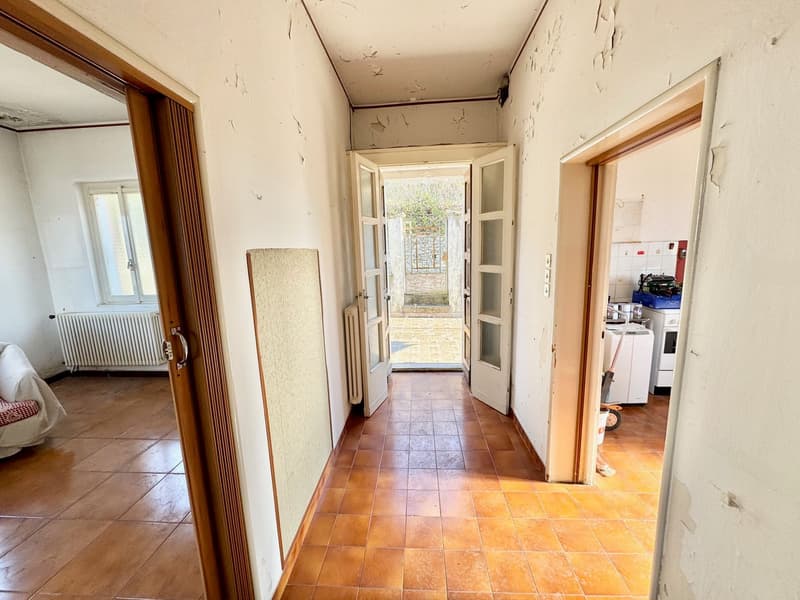 The image shows the interior of a residential property. The hallway has a tiled floor and leads to several rooms, including what appears to be a living room and a kitchen. The walls are in need of some repair, with visible cracks and peeling paint. The do