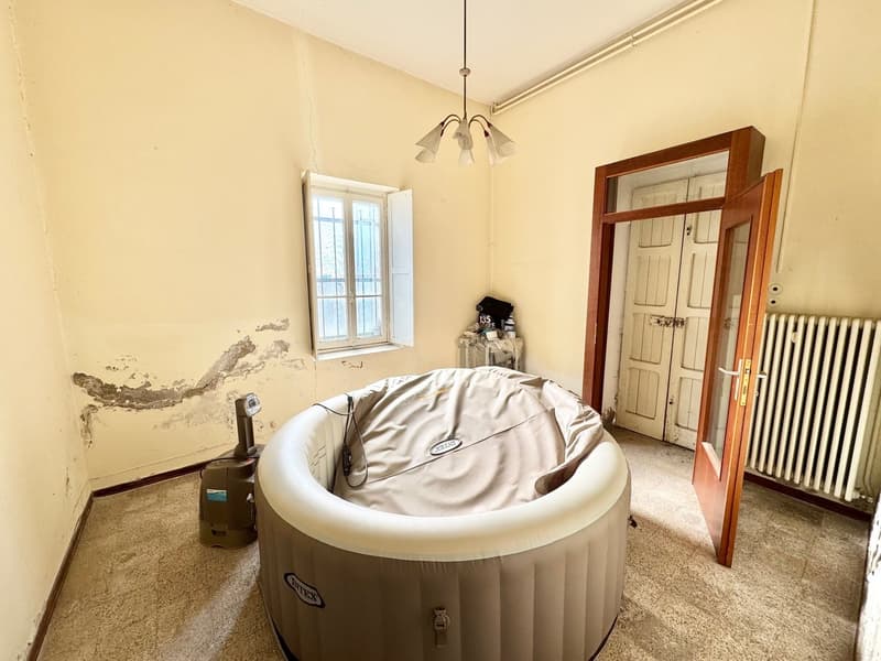 This image shows a room with a white inflatable hot tub or spa in the center. The room has yellow walls, a window, and a wooden door. There is a chandelier-style light fixture hanging from the ceiling. The floor appears to be carpeted. There is some damag