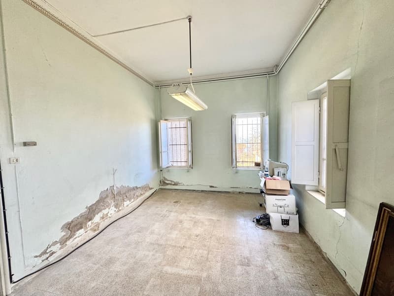 This image shows a room that appears to be in need of renovation. The walls have peeling paint and the floor is bare concrete. There are two windows that provide natural light, and a light fixture hanging from the ceiling. The room also contains some boxe