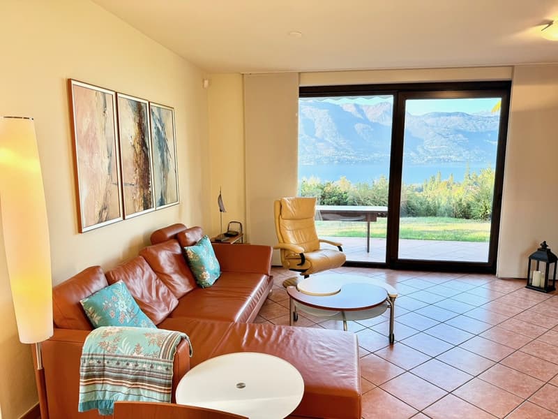 Spacious living room with large windows offering a scenic view of the mountains and surrounding landscape. The room features a leather sofa and armchair, a coffee table, and framed artwork on the walls. The floor is tiled, and the overall decor has a warm