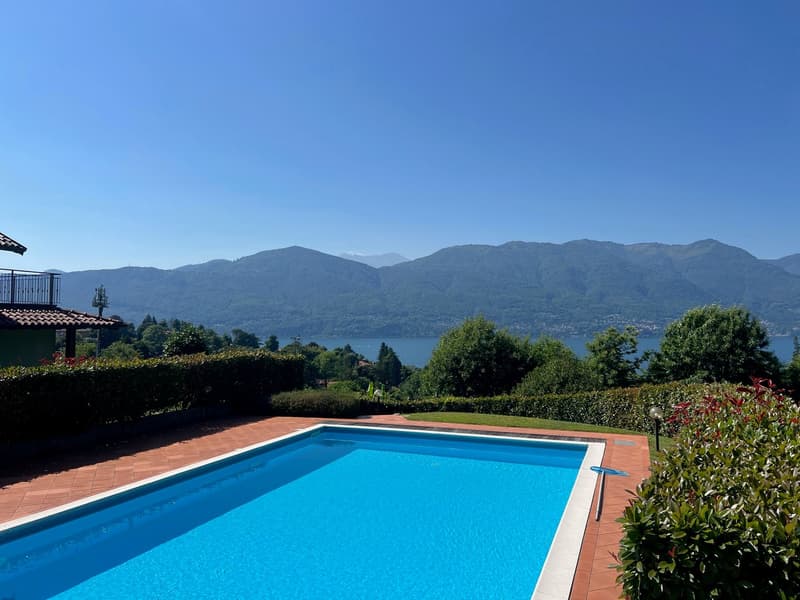 Outdoor swimming pool with clear blue water, surrounded by lush greenery and trees. In the background, there are mountains and a lake, providing a scenic view.