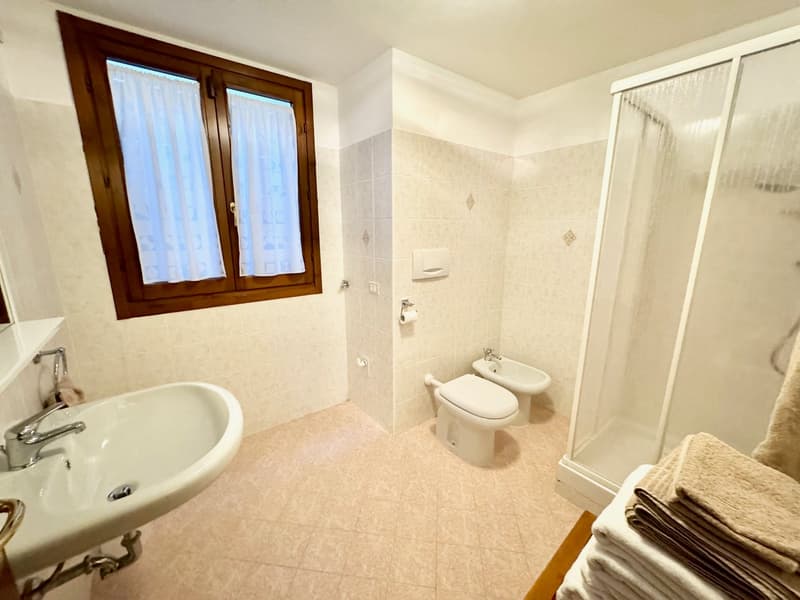 Spacious bathroom with beige tile flooring, a sink, toilet, and a shower enclosure with a glass door. The room has a wooden framed window providing natural light.