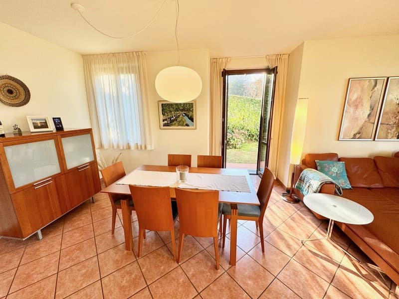 Spacious dining area with wooden table and chairs, wooden cabinets, large windows overlooking a garden, and artwork on the walls.