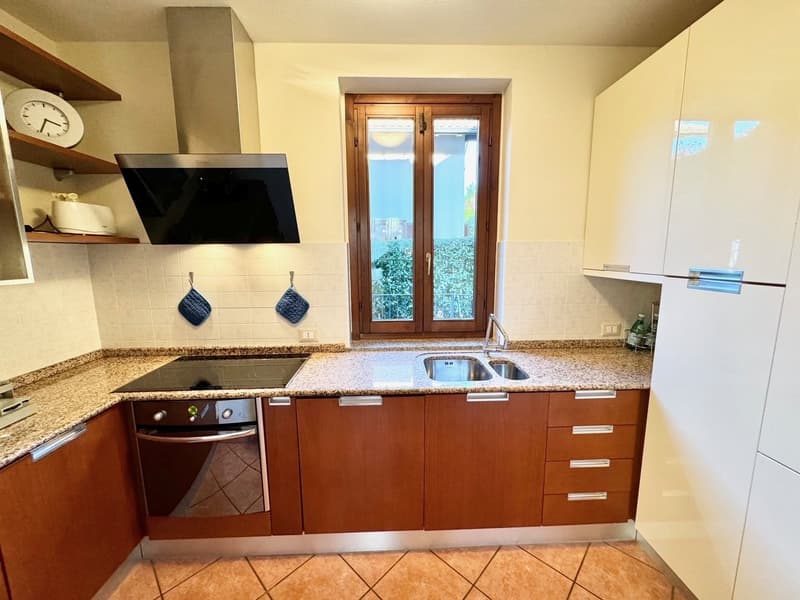 The image shows a modern kitchen with wooden cabinets, granite countertops, a double sink, and a dishwasher. There is a TV mounted on the wall and a sliding glass door leading to an outdoor area.
