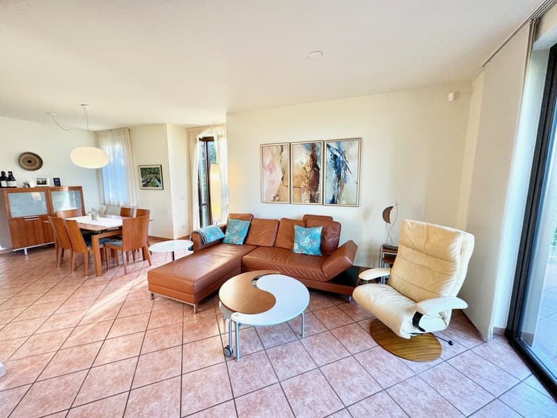 Spacious open-plan living area with hardwood floors, beige walls, large windows, leather sofa, armchair, coffee table, and dining table with chairs. The room has a modern and cozy feel with artwork on the walls.