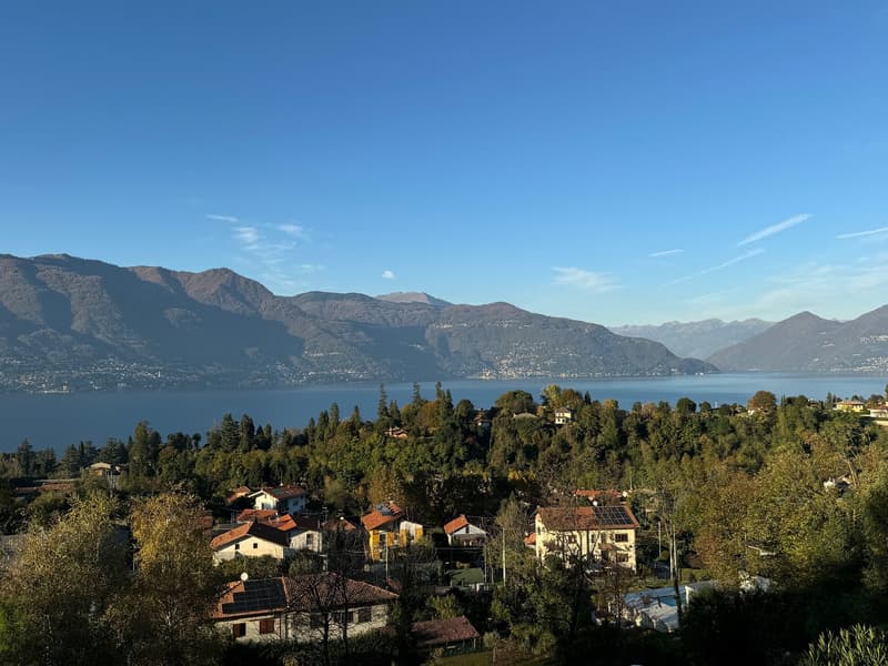 Scenic view of a lakeside town surrounded by mountains. The image shows a picturesque landscape with a large body of water, likely a lake, surrounded by lush green hills and mountains in the background. The town is composed of colorful houses and building