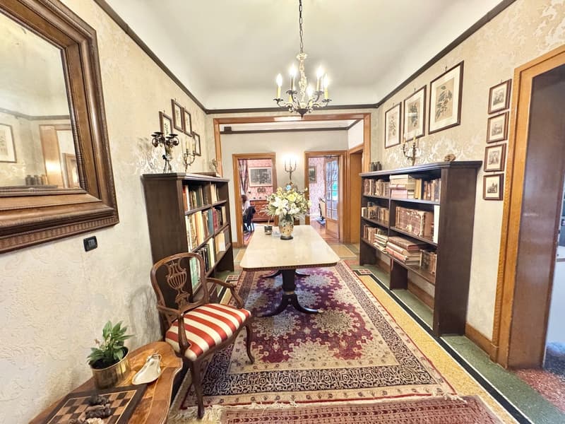 Spacious dining room with ornate wooden furniture, including a large dining table, bookshelves, and a chandelier. The room features a patterned rug, wall decor, and a fireplace.