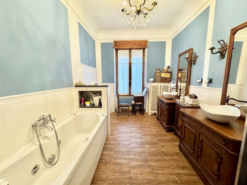 Spacious bathroom with a clawfoot bathtub, wooden vanity with vessel sink, and a fireplace. The room has blue walls, hardwood flooring, and a crystal chandelier.