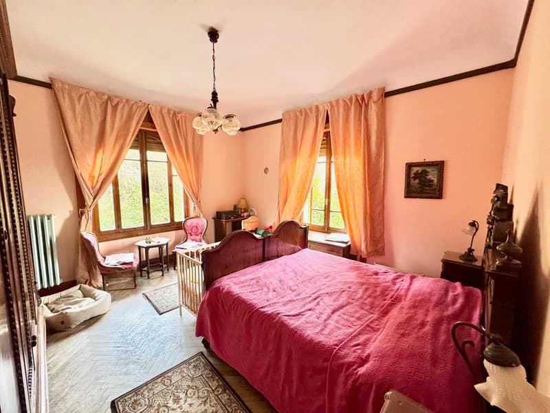 Large bedroom with wooden furniture, including a bed with a red comforter, a dresser, and a chair. The room has large windows with curtains, and a chandelier hanging from the ceiling.