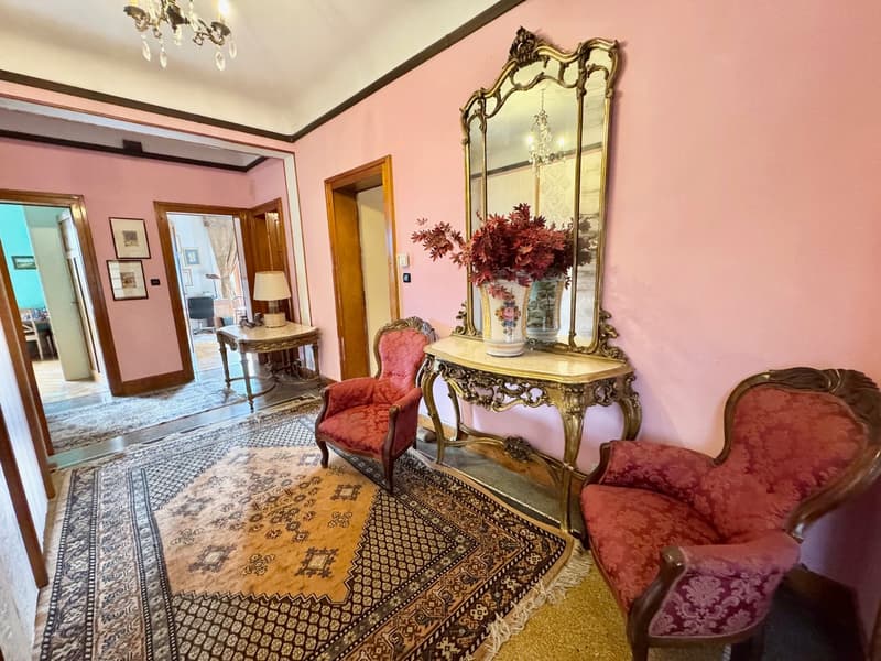 Ornate and luxurious living room with pink walls, antique furniture, a large ornate mirror, and a vase of red flowers. The room features a patterned rug, a chandelier, and a fireplace.