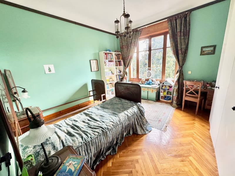 Spacious bedroom with mint green walls, hardwood flooring, large window with curtains, antique-style chandelier, wooden bed frame with patterned bedding, bookshelf, and storage trunk.