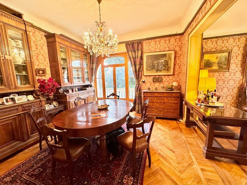 Ornate wooden furniture, including a large dining table with chairs, a china cabinet, and a dresser. The room has patterned wallpaper, a crystal chandelier, and a large window with curtains. There is also a fireplace visible.