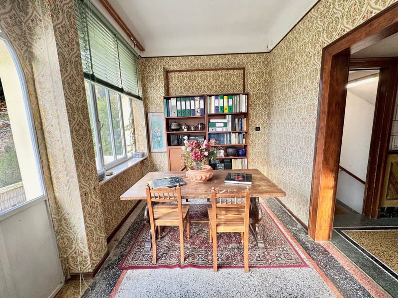 Spacious dining room with patterned wallpaper, wooden dining table and chairs, built-in bookshelf, and large window overlooking a balcony or terrace