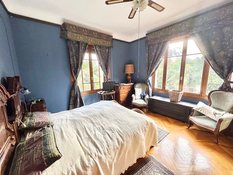 Large bedroom with blue walls, wooden floors, large windows overlooking a garden, antique furniture including a dresser and armchair, and patterned curtains and bedding.
