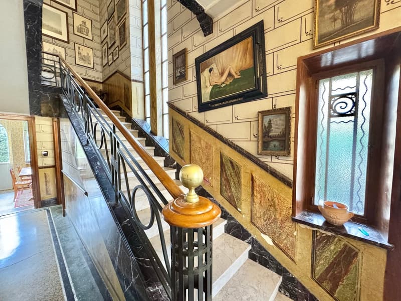 Ornate wrought-iron staircase railing, wooden handrail, marble steps, framed artwork and decor on the walls, antique-style light fixture