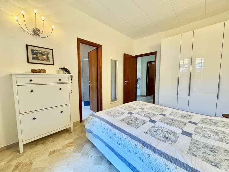 Spacious bedroom with white walls, hardwood floors, a wooden dresser, and a large bed with a patterned comforter. The room has multiple doors, suggesting access to a balcony or terrace.