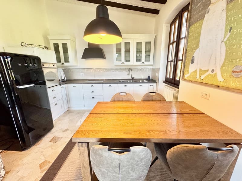 The image shows a combined kitchen and dining area with white cabinets, a large wooden dining table, and a black Smeg refrigerator. The kitchen features a tile backsplash and a large pendant light fixture. The dining area has a wooden table and gray uphol