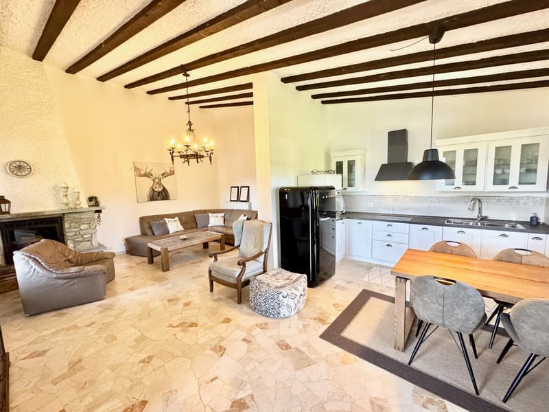 The image shows an open-plan living area with a living room, dining area, and kitchen. The room has exposed wooden beams on the ceiling, a stone fireplace, and a mix of modern and rustic furnishings. The kitchen features white cabinets, black appliances,