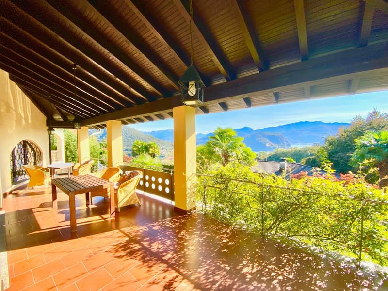 A covered outdoor area with wooden flooring and ceiling, a wooden table and chairs, a lamp, and a fence with a view of mountains and trees.