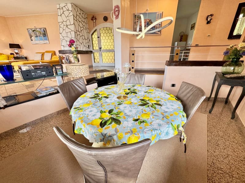 Dining table with a colorful tablecloth, 4 chairs, a shelf with books, a lamp, flowers, a cabinet, carpet, a kitchen in the background