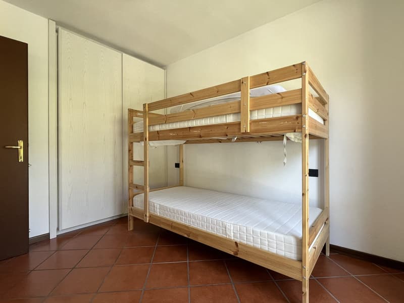 double bunk bed with white mattresses, wooden frame, brown tile floor, white wall, wooden wardrobe, door on the left
