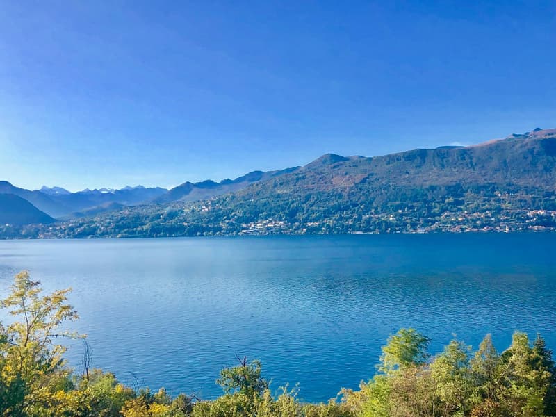Lake surrounded by mountains, clear blue water, greenery, distant buildings