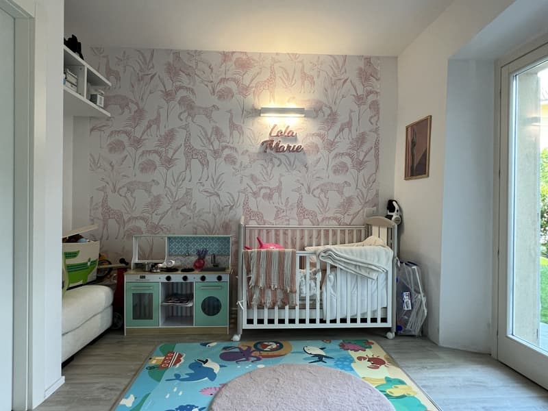 Children's room with wallpaper of animals, crib, toy kitchen, rug, shelves, and a desk with a lamp
