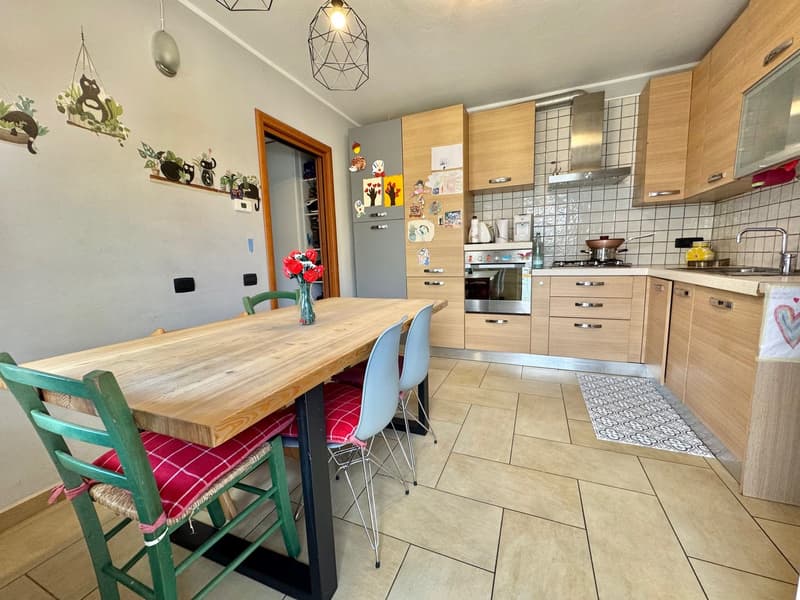 Spacious kitchen with wooden cabinets, stainless steel appliances, tiled floor, dining table with colorful chairs, decorative wall shelves, and a vase of red flowers on the table