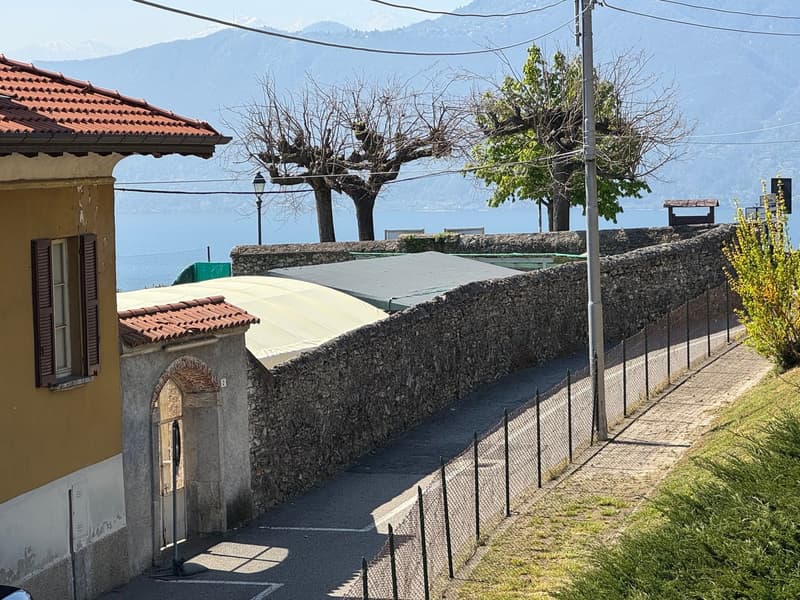 The image shows a residential area with a stone wall, a paved walkway, and several buildings with tiled roofs. There are trees and a parking area visible. The surrounding landscape includes mountains in the background, indicating a scenic, rural setting.