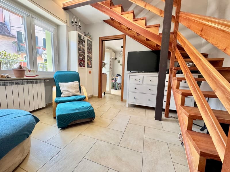 Cozy living room with wooden staircase, teal armchair, dresser, and TV stand. Large windows provide natural light and a view of the balcony.