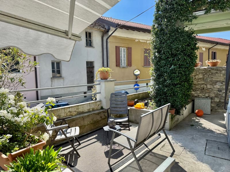 Two-story house with yellow exterior, red tile roof, and green foliage. Outdoor seating area with chairs and table on a paved terrace. Potted plants and flowers around the property. Appears to have a parking space or driveway.