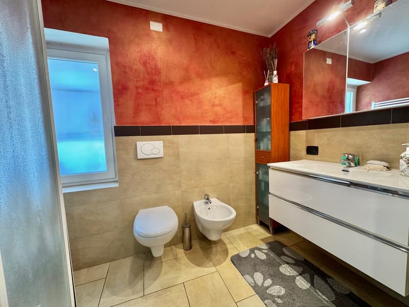 The image shows a bathroom with orange-colored walls, beige tile flooring, a white toilet, a bidet, and a vanity with a sink and drawers. There is a window that provides natural light to the space.
