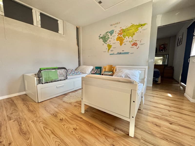 Spacious bedroom with hardwood floors, white walls, and a large world map mural. The room contains two twin beds, a dresser with drawers, and a chair.