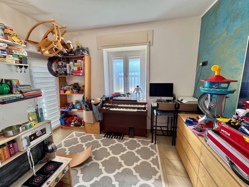 Cluttered bedroom with a piano, shelves filled with toys and books, a TV, and a window. The room has a patterned rug on the floor.