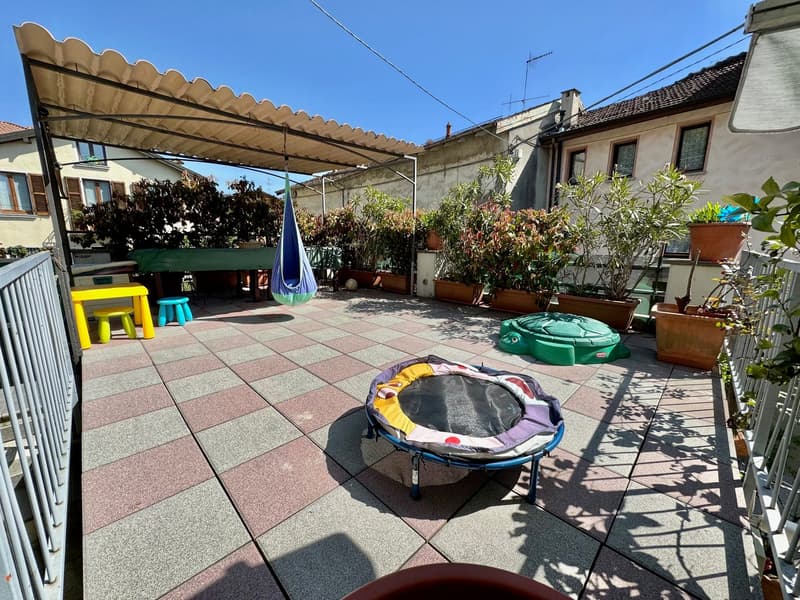Paved outdoor terrace with potted plants, a trampoline, and colorful furniture. There is a covered seating area and a parking space visible.