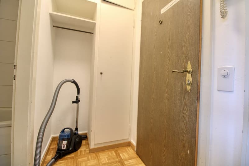 A small room with a wooden floor, a white door, a white closet, a white wall, a switch, and a vacuum cleaner.