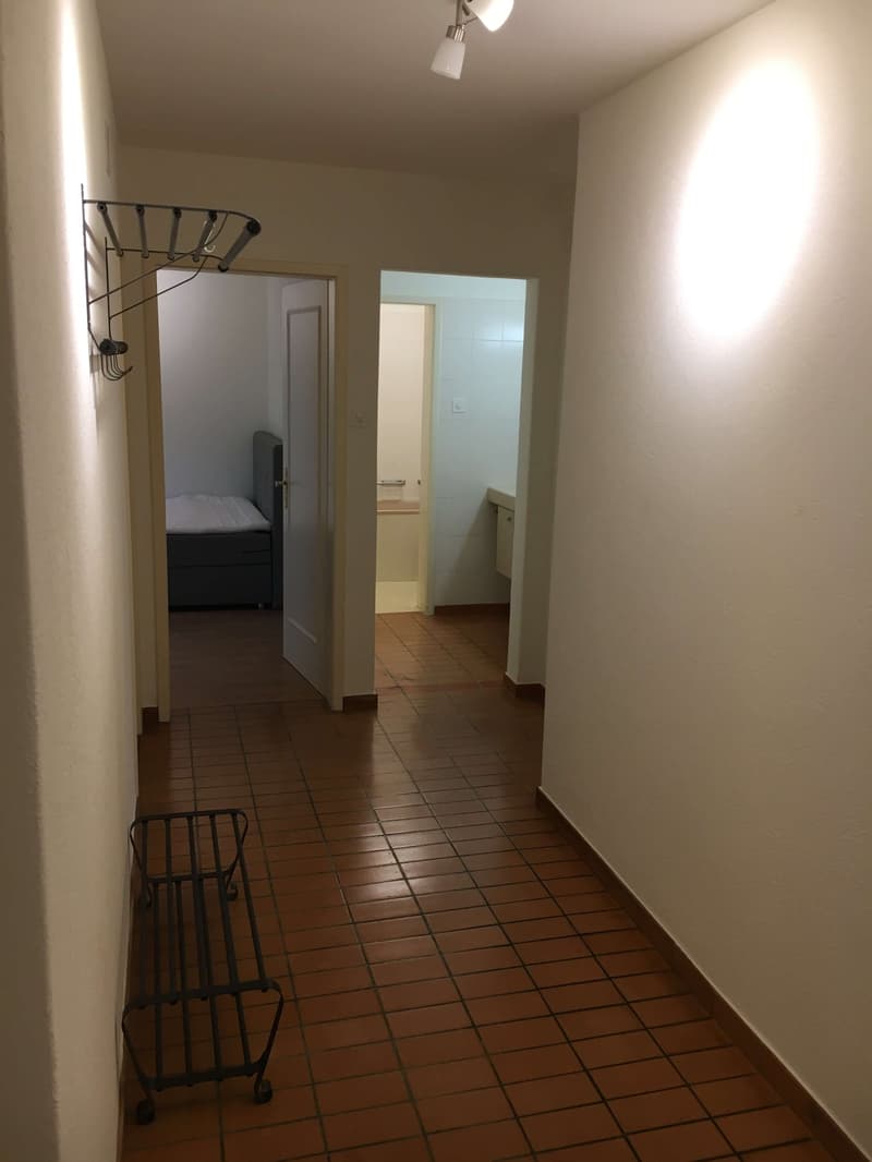 A hallway with brown tiles, an open door leading to a bedroom with a bed, another door leading to a bathroom, a small bench on wheels, and ceiling lamps