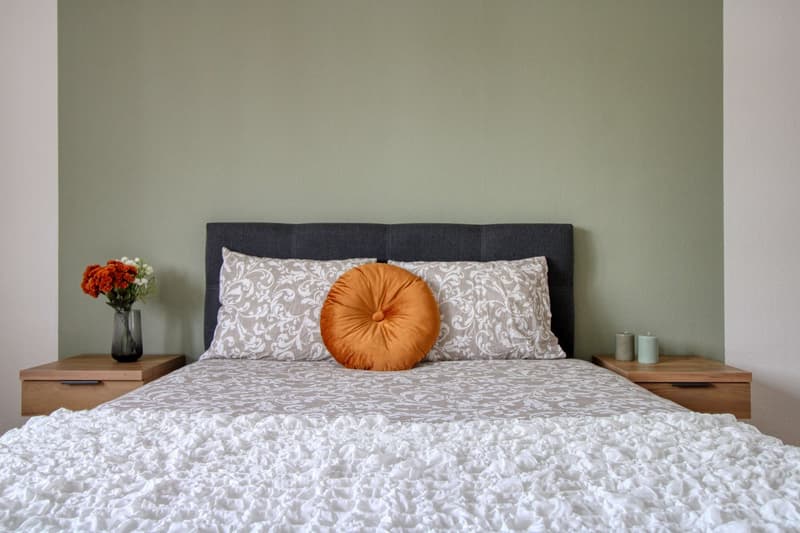 A neatly made bed with a white bedspread, two nightstands with candles, a vase with flowers, and a gray headboard.