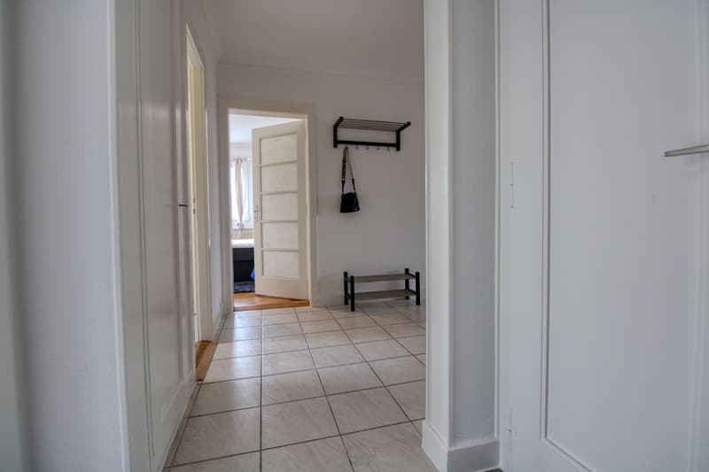 Empty hallway with white walls, tiled floor, white doors, open door, small bench, bag hanging on wall