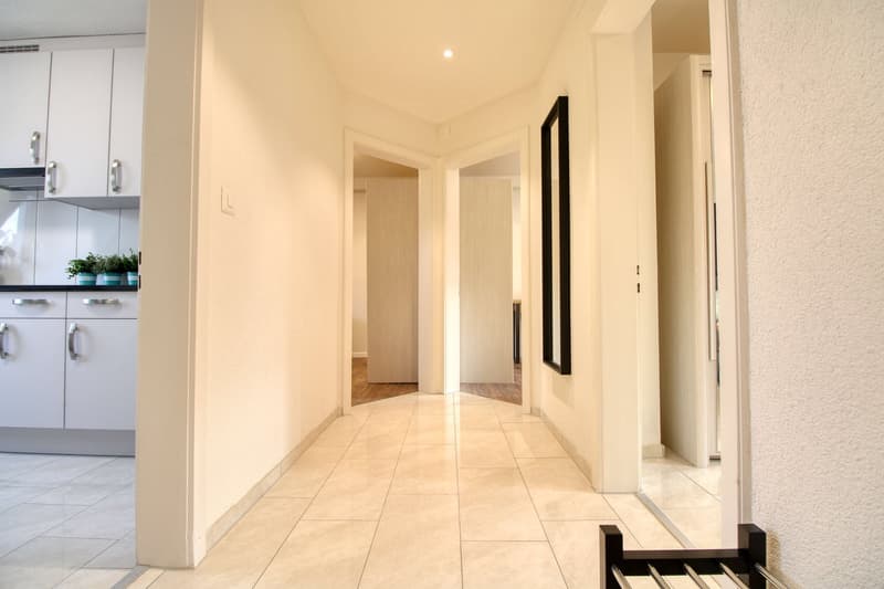 wide hallway, cream-colored tiles, open doors, modern kitchen cabinets, potted plants