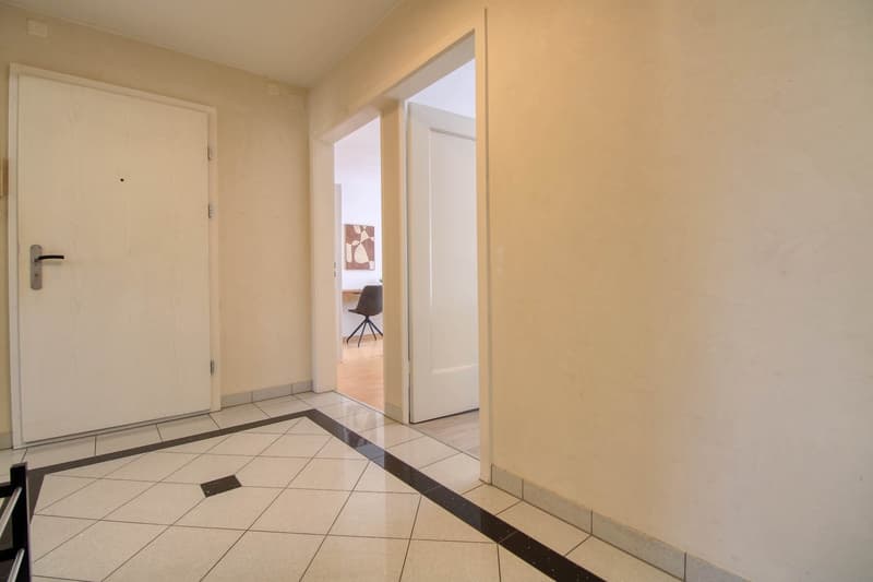 The image shows the entrance of an apartment. The floor is tiled with a pattern of light and dark tiles. The walls are painted in a neutral beige color. There is a white door leading into the apartment, and another doorway visible in the background.