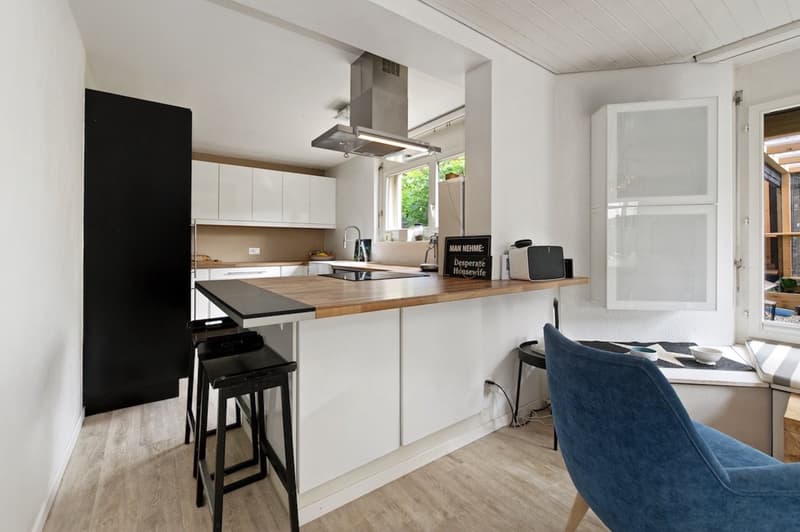 open-plan kitchen with wooden countertops, black stools, blue chair, modern appliances, large windows, wooden flooring, white cabinets