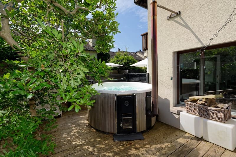 hot tub with water, wicker basket with firewood, stone seat, black step, white container, wooden deck, trees