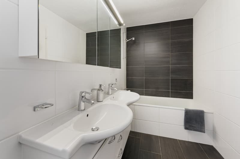 Bathroom: white tiled walls, white bathtub, white double sink, mirror above sink, gray towel on side of bathtub.