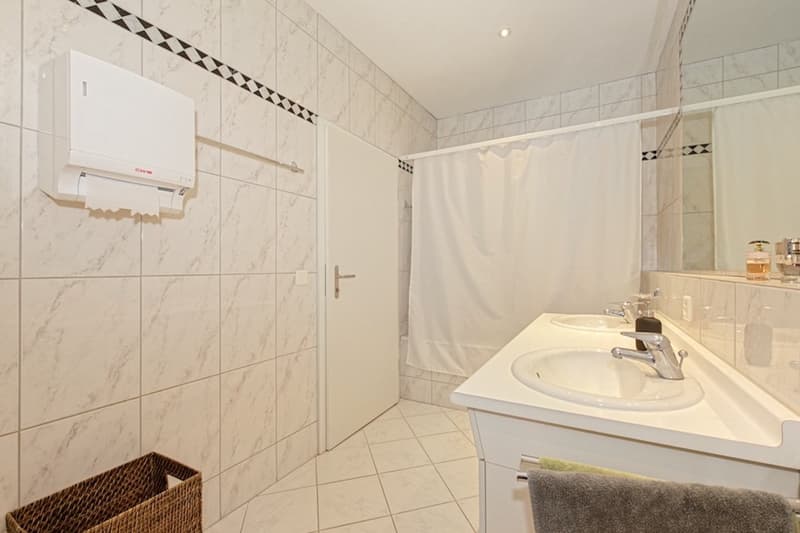 tiled walls, double sink, white fixtures, white door, towel, small basket, mirror, roll paper towel holder