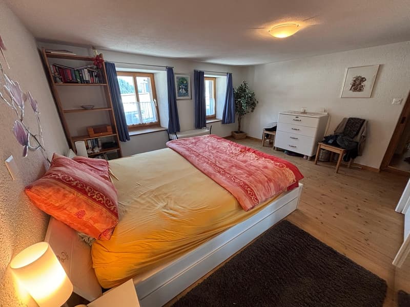 a bed with a yellow bedspread, a white headboard, and two orange pillows with red and gold designs. On the left side of the bed is a lamp, and on the right is a shelf with books and a small box. There is a picture frame hanging on the wall, and a wooden c