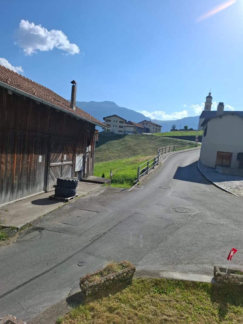 2 story brown wooden house, red tile roof, gable end facing street, large double doors, white walled house on right, mountain in background, red flag