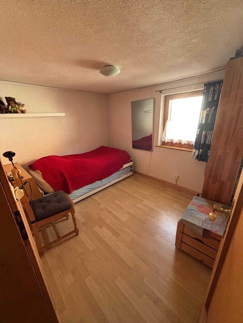 small room with a bed, wooden floor, white walls, ceiling light, small chair, window with white curtains, shelf, wardrobe, small table with lamp