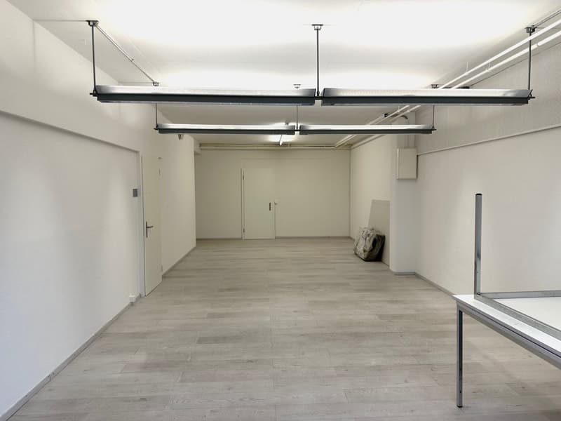 Large open space with concrete flooring, exposed ceiling with hanging lighting fixtures, and a single door leading to another room or area.